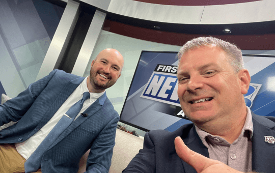 two males on a television news set with one holding a thumbs up